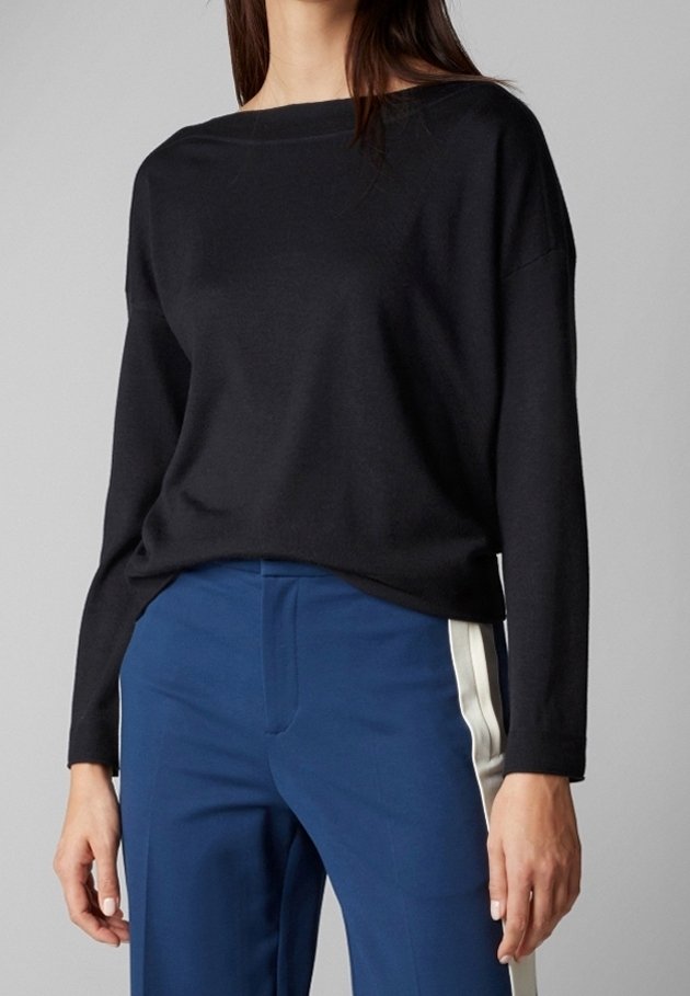 Femme portant un haut noir à manches longues et un pantalon bleu avec des bandes blanches sur les côtés, debout devant un fond gris uni.