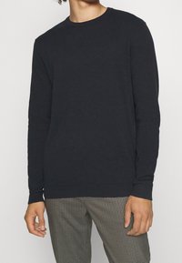 Maglione blu scuro a girocollo realizzato in morbido tessuto a maglia, con polsini e orlo a coste, abbinato a pantaloni a quadri. Nessun motivo particolare sul maglione.