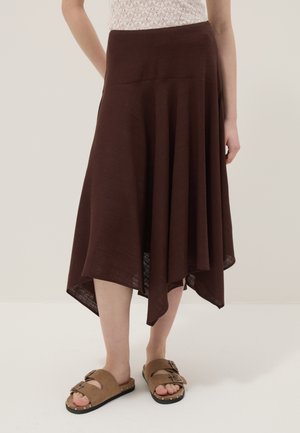 Mujer vistiendo una falda midi asimétrica marrón oscuro, blusa blanca de encaje y sandalias beige con hebilla, de pie sobre un fondo liso y claro.