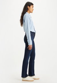 Chemise boutonnée bleu clair à manches longues, associée à un jean foncé évasé et des baskets blanches. Design minimaliste et texture lisse.