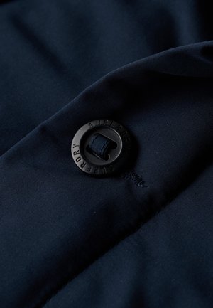 Dark navy fabric with a smooth texture featuring a black round button labeled "SUPERDRY" and a small loop of elastic.