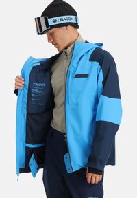 Blau und marineblaue wasserdichte Skijacke mit einem durchgängigen Reißverschluss, verstellbaren Bündchen und einer inneren Mesh-Tasche. Enthält Spyder-Branding innen.