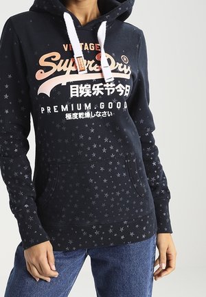 Granatowa bluza z kapturem w białe gwiazdki, białe sznurki i wielokolorowe logo "SuperDry" z tekstem japońskim i angielskim, noszona z niebieskimi dżinsami.