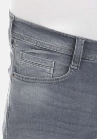 Zbliżenie na osobę noszącą jasnoszare dżinsy z widoczną pętlą na pasek i przednią kieszenią, oraz białą koszulę włożoną w spodnie.