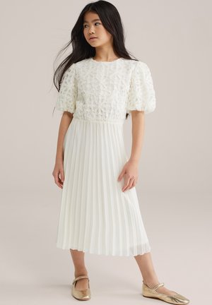 Balta kleita ar reljefu ziedu rakstu uz īsām pumpuru piedurknēm, krokotu svārku un apaviem zelta krāsas Mary Jane stilā. Gluda auduma apdare.