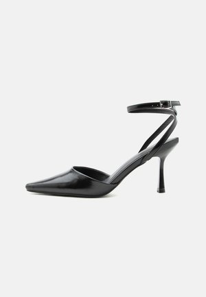 Schwarzer spitzer High Heel mit dünnen Knöchelriemen und silberner Schnalle auf einfachem hellem Hintergrund.