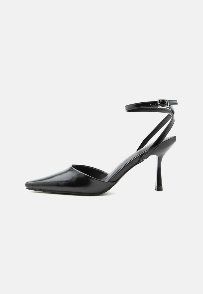 Μαύρη γόβα μυτερή με λεπτά λουράκια στον αστράγαλο και ασημένια πόρπη σε απλό ανοιχτό φόντο.