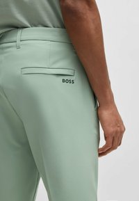 Jasnozielone spodnie o gładkiej fakturze, z tylną kieszenią z klapką oraz nazwą marki "BOSS" wydrukowaną na czarno z boku.