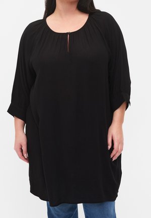 Frau trägt lockere schwarze Tunika mit 3/4-Ärmeln, Knopf am Schlitz am Ausschnitt und blaue Jeans, steht vor einfachem Hintergrund.