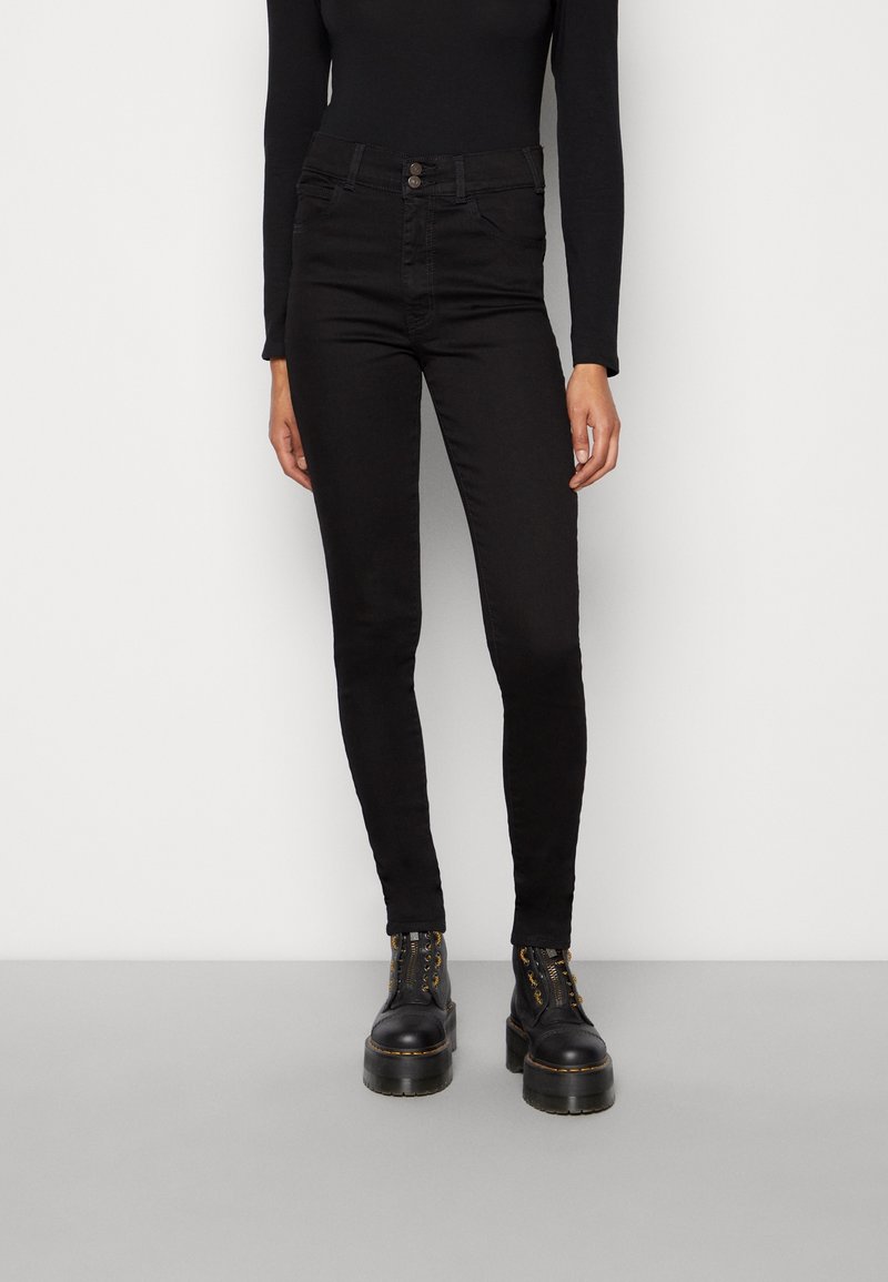 Personne portant un pantalon noir ajusté taille haute, un haut noir à manches longues et des bottes à plateforme épaisses noires, debout devant un fond blanc.
