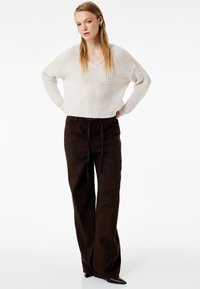 Ung kvinde med langt blond hår iført en hvid strikket sweater, mørkebrun bukser med vide ben og bælte, samt sorte spidse sko og stående.