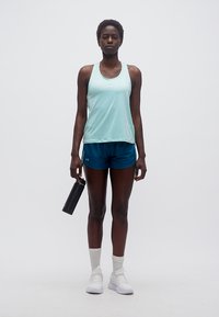 Femme en tenue de sport tenant une bouteille d'eau noire, debout devant un fond clair uni.