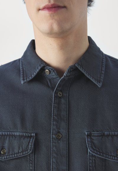 Mørkeblå denimskjorte med spiss krage, to brystlommer og metallknapper. Stoffet har en teksturert overflate.