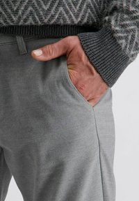 Hellegraue Baumwollmischhose mit glatter Textur, die seitliche Taschen und eine lockere Passform aufweist. Eine Hand ruht im linken Taschenbereich.
