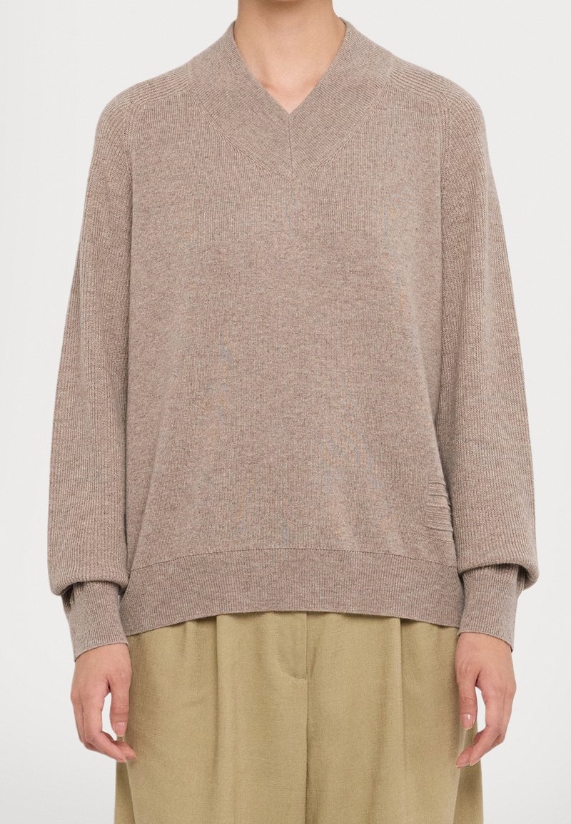 Personne portant un pull beige en maille à col en V avec des manches longues et des poignets côtelés, assorti à un pantalon ample de couleur sable.