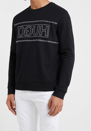 Mand iført sort langærmet sweatshirt med inverteret hvidt "HUGO" logo og hvide bukser, stående mod ensfarvet lyst baggrund.