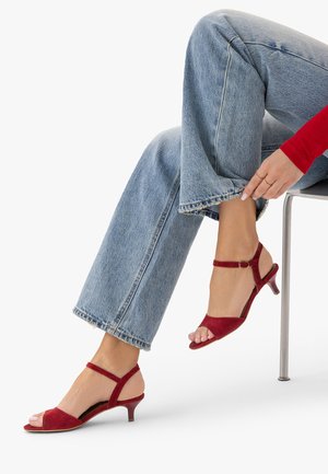 Person trägt hellblaue Jeans und rote Sandalen mit offenem Zehenbereich und kleinem Absatz, sitzt und trägt den Jeanssaum mit der Hand hoch.