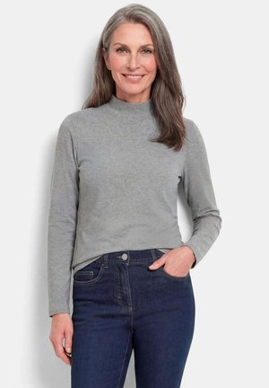 Lächelnde Frau mit langen grauen Haaren, die ein graues Langarmshirt und dunkelblaue Jeans trägt und mit einer Hand in der Tasche steht.