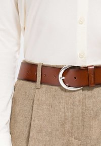 Cintura in pelle marrone con una fibbia rotonda in argento indossata sopra pantaloni beige strutturati e una camicia bianca con bottoni.