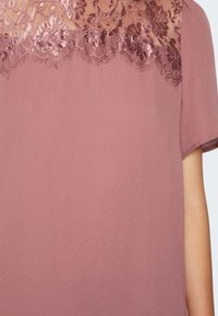 Krátký rukáv top v jemné růžové barvě s krajkovými detaily na výstřihu, zdobený květinovými vzory a hladkou texturou látky.