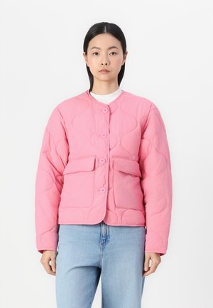 Woman wearing a quilted pink buttoned jacket with large flap pockets, white top underneath, and light blue jeans, standing against a white background.