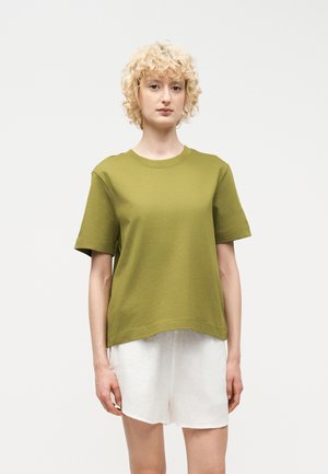 Ξανθό άτομο με κοντά, σγουρά μαλλιά που φοράει ελιάς πράσινο κοντομάνικο πουκάμισο και λευκό σορτς, στέκεται μπροστά σε απλό φόντο.