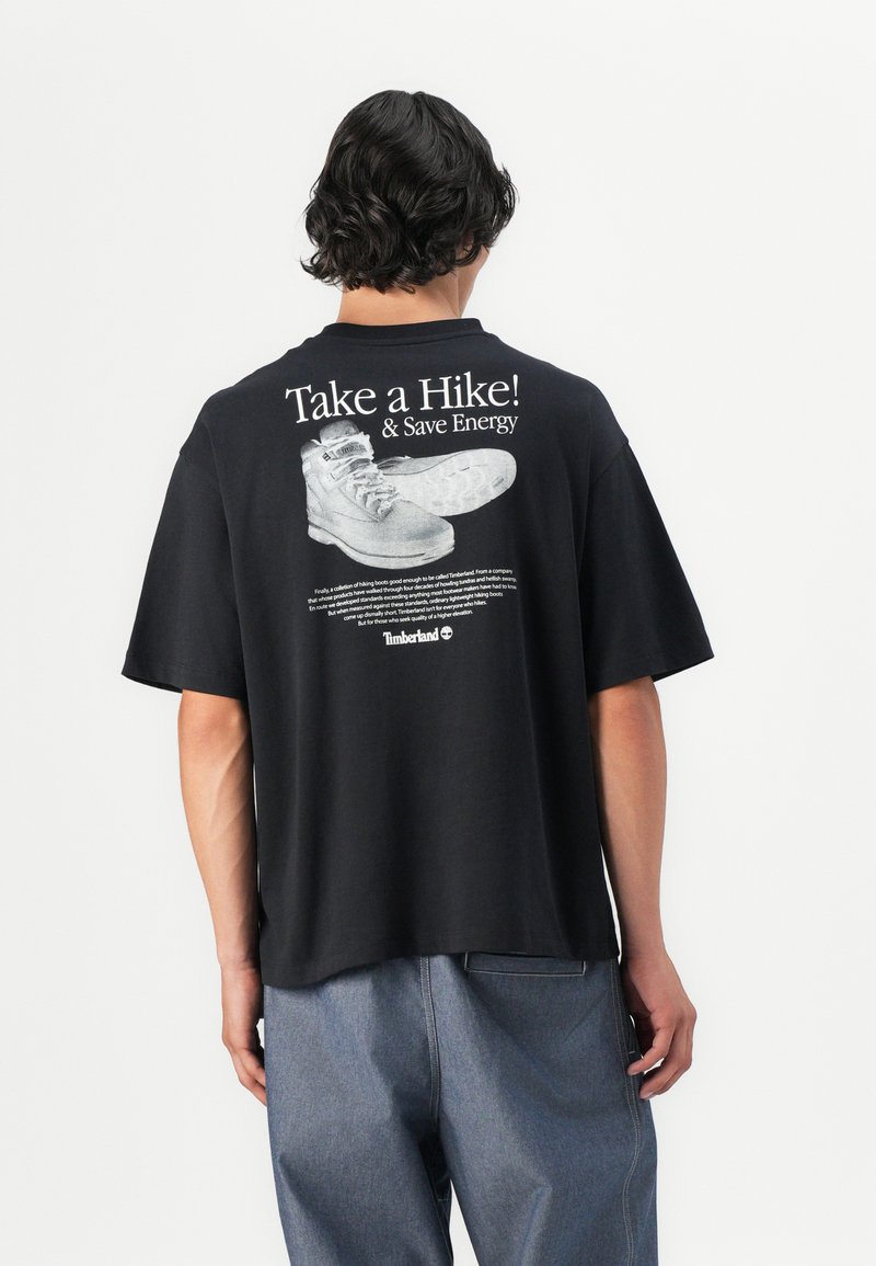 T-shirt nera oversize con una stampa grafica di stivali e testo sul retro. Maniche corte, vestibilità rilassata e materiale in cotone.