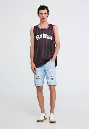 Maglietta nera senza maniche con strisce rosse e testo "SAN DIEGO", abbinata a pantaloni corti lion blu chiaro strappati e sneakers bianche con dettagli neri.