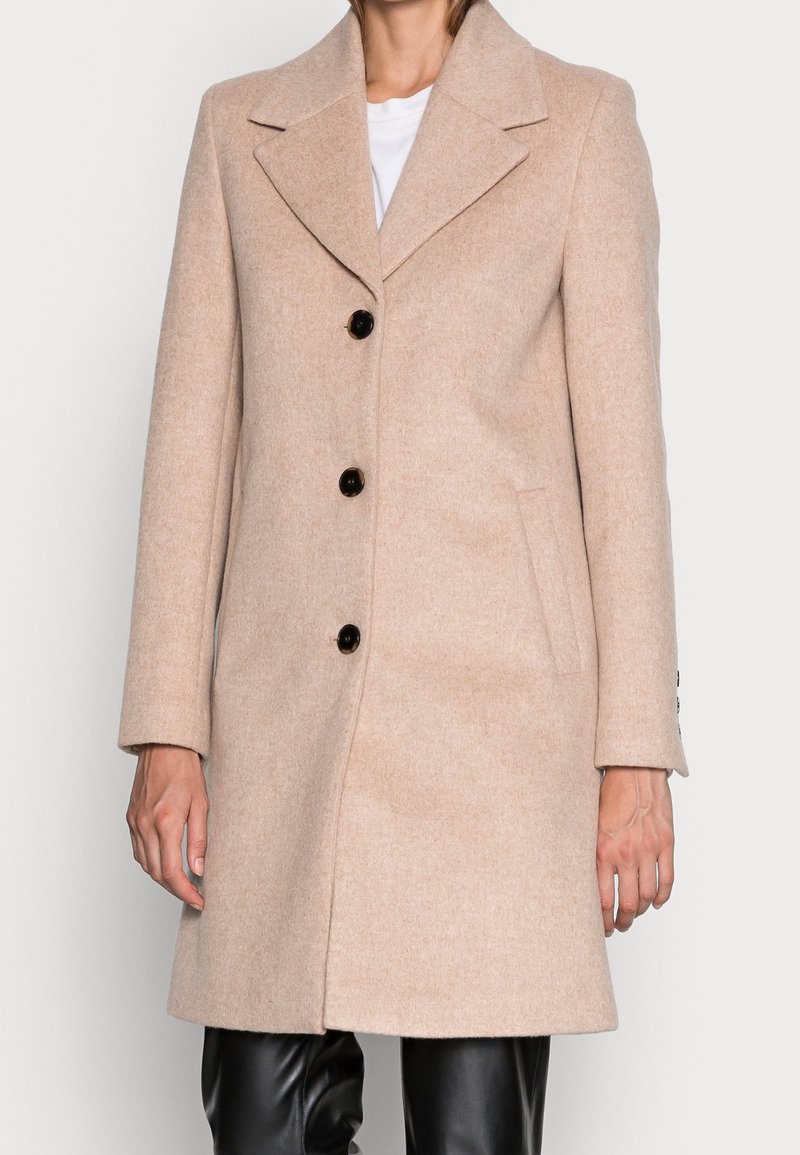 Femme portant un manteau en laine beige avec des boutons noirs sur une chemise blanche et un pantalon en cuir noir, se tenant devant un fond uni.