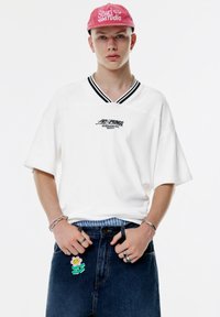 Ung person iført en rød kasket, en hvid oversized skjorte med sort tekst, blå jeans og ringe, armbånd, med en blomst vedhæng på jeans.