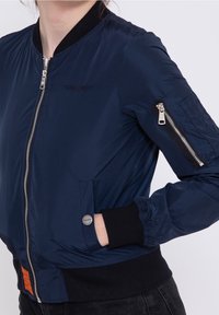 Veste bomber bleu marine avec fermeture éclair à l'avant, poignets et ourlet côtelés, poches latérales et une poche zippée sur la manche avec un détail de logo.