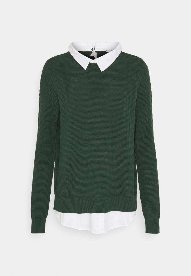 Pull en maille texturée vert foncé superposé à une chemise blanche à col visible avec col et ourlet apparents sur un fond uni.