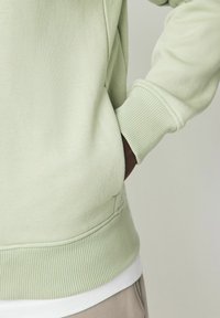 Világoszöld pulóver, puha anyagból készült, bordás mandzsettákkal és szegéllyel. Egy fehér réteg látható alatta. Az anyag sima textúrájú.