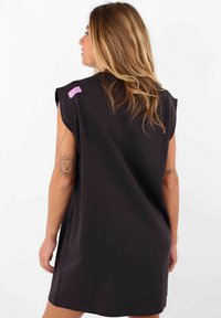 Robe noire oversize à manches courtes, avec un accent rose à l'épaule. Texture lisse, design à mi-genoux, coupe décontractée.