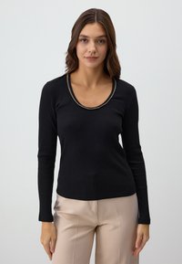 Zwarte geribbelde top met lange mouwen, voorzien van een ronde halslijn met een gouden kettingaccent. Gedragen over beige broeken, die een aansluitende silhouet benadrukken.