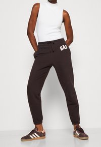 Pantalones deportivos marrón oscuro con cintura ajustable, con el logo blanco "GAP" en la cadera izquierda, combinados con zapatillas oscuras con rayas blancas.