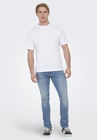 T-shirt bianca a maniche corte abbinata a jeans fitted azzurri chiari e sneakers in bianco e nero. L'aspetto complessivo è casual e semplice.