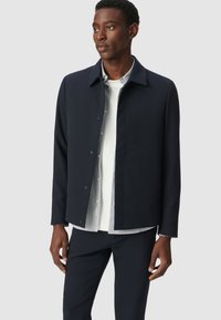 Chaqueta navy con un cuello estructurado, cierre frontal a presión y mangas largas, superpuesta sobre una camisa blanca con rayas azules, combinada con pantalones oscuros.