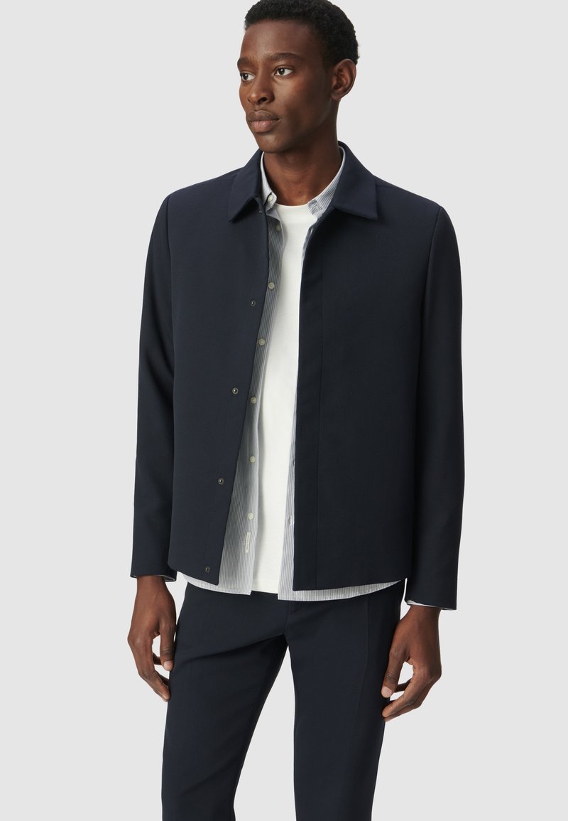 Chaqueta navy con un cuello estructurado, cierre frontal a presión y mangas largas, superpuesta sobre una camisa blanca con rayas azules, combinada con pantalones oscuros.