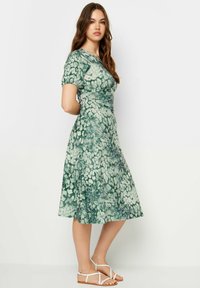 Blumenmuster-Kleid in Grüntönen, knielang, mit kurzen Puffärmeln, tailliert, fließender Rock, kombiniert mit hellbraunen flachen Sandalen.