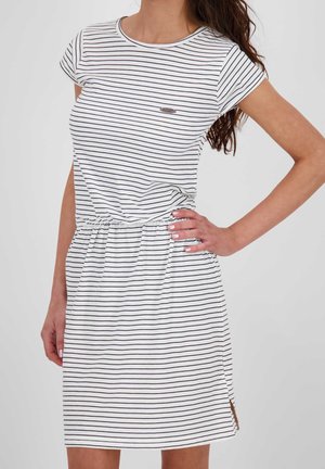 Kobieta w białej sukience z krótkim rękawem w cienkie czarne poziome paski, podkreślonej w talii, z ręką opartą na biodrze.