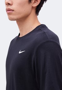Jeune personne portant un t-shirt noir à manches longues avec un petit logo blanc Nike sur la poitrine, vu de profil et recadré au niveau du cou.