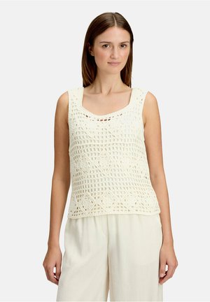 Femme portant un top blanc en crochet sans manches et un pantalon blanc ample, debout devant un fond uni clair.