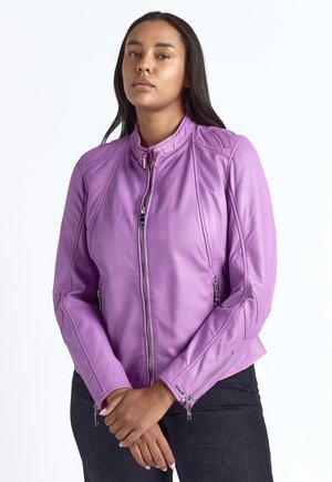 Vrouw met lang donker haar, die een paarse leren jas met rits en een zwarte broek draagt, staat met de handen voor zich geklemd.