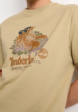 T-shirt beige avec un graphique de bottes de randonnée brunes sur de la mousse verte et violette, avec le texte "Timberland BOOTS VTS."