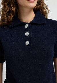 Kvinde iført en mørkeblå glitrende strik polo t-shirt med krave og tre dekorative runde perleknapper fortil.