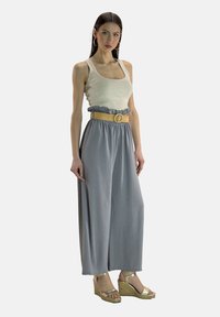 Pantalon large gris clair avec une taille froncée, associé à un débardeur beige. Sandales à talons compensés dorées et une ceinture beige avec une boucle circulaire en accent.