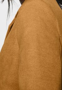 Tan wool blend jacket featuring a textured finish, structured shoulders, and visible stitching along the seams.
