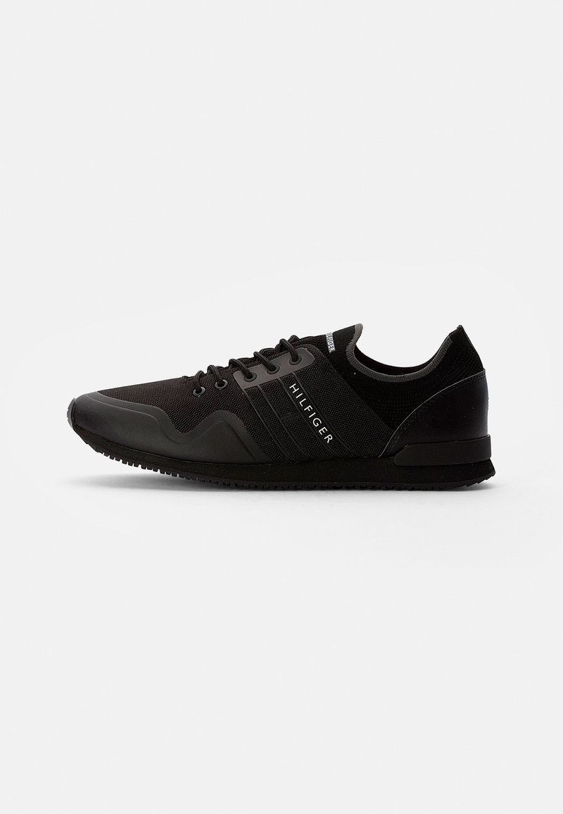 Chaussure de sport basse noire avec lacets à l'avant, semelle texturée et texte "HILFIGER" sur le panneau latéral.
