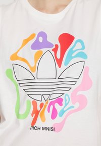 Wit T-shirt met kleurrijke abstracte vormen en de tekst "LOVE UNITE" boven een zwart Adidas-logo. Onderste text bevat "RICH MNISI".